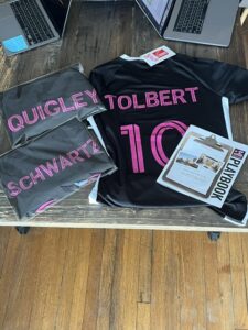 black football jerseys with pink names quigley, tolbert and schwartz and the number 10, laid on a wooden table alongside a playbook and laptops.