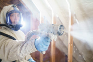 worker in full protective suit and respirator spraying a nozzle in a dusty attic.