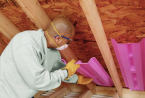man wearing a mask and gloves cuts pink insulation panels between wooden rafters in an attic.