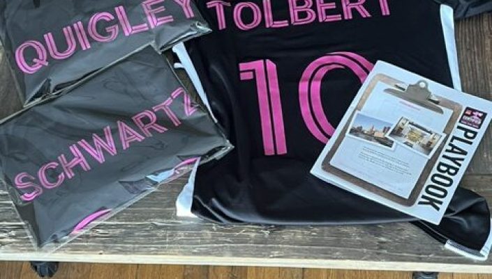 Black football jerseys with pink names Quigley, Tolbert and Schwartz and the number 10, laid on a wooden table alongside a Playbook and laptops.