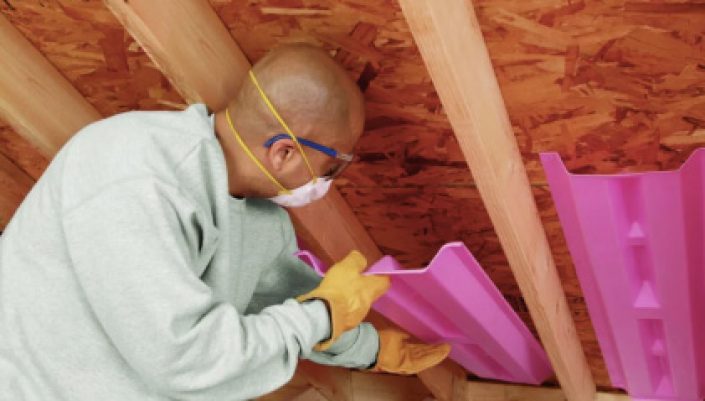 Man wearing a mask and gloves cuts pink insulation panels between wooden rafters in an attic.
